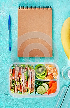 School lunch box with sandwich, vegetables, water, and fruits on table.