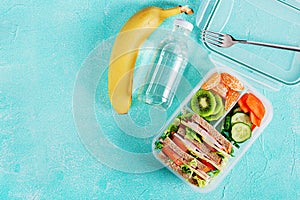 School lunch box with sandwich, vegetables, water, and fruits on table.