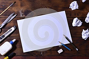 A school or office still life with a white blank sheet of paper and many office supplies. The school supplies lie on a brown wood