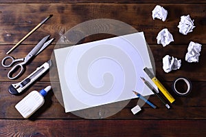 A school or office still life with a white blank sheet of paper and many office supplies. The school supplies lie on a brown wood