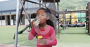 In a school playground, a young biracial girl is blowing bubbles