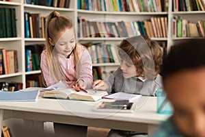 School pupils reading book together in library, doing homework or preparing for classes, sitting at desk and chatting