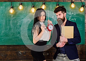 School rules concept. Man with beard hold book and girl teacher holds alarm clock, chalkboard on background. Lady