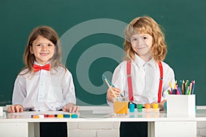 School smiling children girl and boy painting with paints color and brush in classroom on school blackboard background