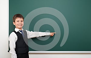School student boy posing at the clean blackboard, show finger and point at, grimacing and emotions, dressed in a black suit, educ