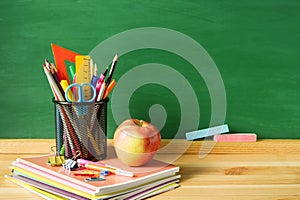 School supplies, pens, pencils in a glass, notebooks, chalk for a blackboard and an apple on a wooden table against the background