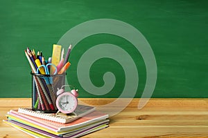 School supplies, pens, pencils, notebooks and an alarm clock on a wooden table against the background of a green school board