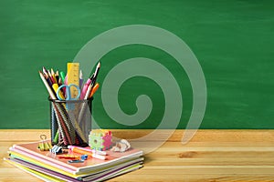 School supplies, pens, pencils, notebooks, sharpener and paper clips on a wooden table against the background of a green school bo