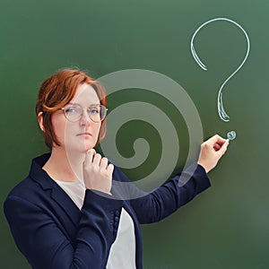 School teacher with a sad face writes in chalk a question mark on a blackboard. Upset woman teacher on green background