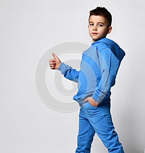 Schoolboy in a blue tracksuit stands sideways and shows a cool sign by raising his thumb up.