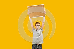 Schoolboy boy raised an empty cork board above his head. Portrait of child in glasses with blank board isolated on yellow