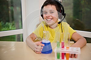 Schoolboy with chemicals in test tubes on tripod, holds flask with blue liquid, completes chemical scientific experiment