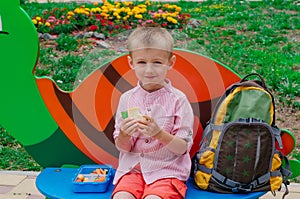 Schoolboy eating his lunch from lunch box sitting on the funny bench outdoor