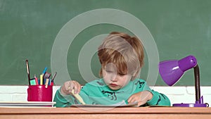 Schoolboy with lesson. Happy smiling pupils drawing at the desk. Kid is learning in class on background of blackboard