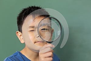 Schoolboy looking through magnifying glass against greenboard in a classroom