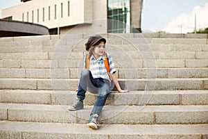 A schoolboy with a paunch on his shoulders and a cap sits on the steps in the city and sadly thinks about something