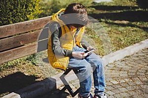 Schoolboy plays game his cell phone sitting on bench in the city park. Boy kid in yellow vest and green hoodie uses