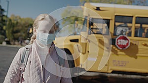 A schoolboy in a protective mask stands in front of a school bus. A Stop sign is visible at the rear. Protecting