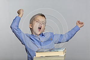A schoolboy sits at a table with books, yawns and sips. Gray background