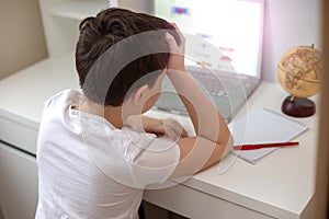 A schoolboy sits at a white computer desk and works at an open laptop.