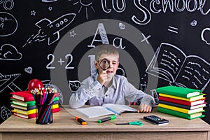 Schoolboy sitting at the desk with books, school supplies, holding with a magnifying glass before the eye. Chalkboard