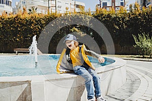 Schoolboy sitting on a fountain in public park during spring sunny day playing with water. Kid in yellow vest chilling