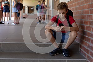 Schoolboy sitting on a step in the schoolyard at elementary school using a smartphone