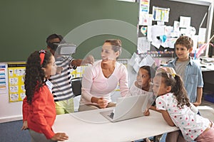 Schoolboy using virtual reality headset at school in classroom with his classmates and her teacher n