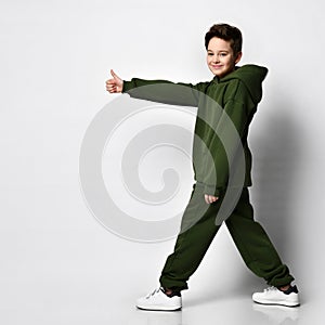 Schoolboy in a warm green tracksuit stands sideways and raising his thumb up.