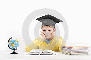 Schoolboy wears the students hat and sitting at desk in class. Portrait of student on white background