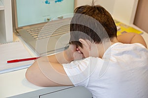 A schoolboy in a white T-shirt, tired of sitting at the monitor with a laptop, holding his head in his hands