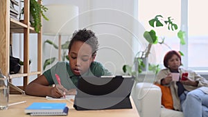 Schoolboy writing and studying in front of tablet screen at table in home room, mom is behind spbd.