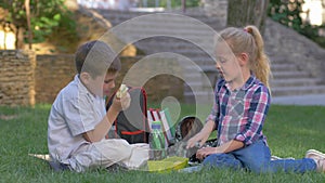 Schoolchildren chat during recess lunch with sandwiches in hands sitting on grass in schoolyard