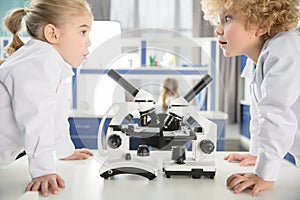 Schoolchildren in lab coats using microscopes and looking at each other