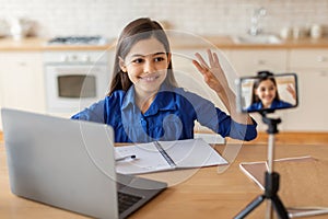 Schooler girl sitting near laptop waving hello to phone indoor