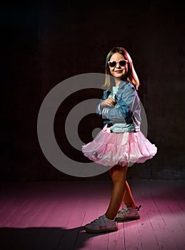 Schoolgirl in blue t-shirt, denim jacket, poofy skirt, sneakers, sunglasses. She smiling, posing on black background. Full length