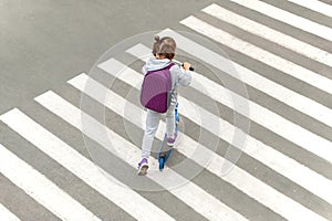 Schoolgirl carrying scooter and crossing road on way to school. Zebra traffic walk way in the city. Pedestrian passing a crosswalk