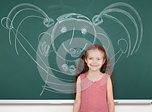 Schoolgirl child in red striped dress drawing happy man on green chalkboard background, summer school vacation concept
