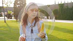 A schoolgirl girl drinks juice and eats a banana in the school yard during a break between lessons.