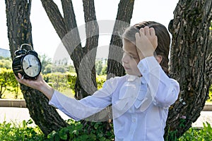 Schoolgirl girl holding an alarm clock in her hand, thinking about time and making faces