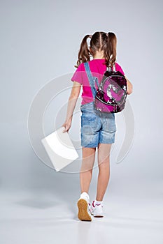 Schoolgirl in glasses with backpack and textbook on the gray background