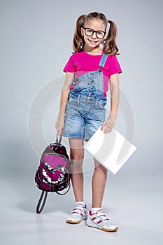 Schoolgirl in glasses with backpack and textbook on the gray background