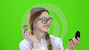 Schoolgirl in glasses powdering her nose with a brush looks in the mirror . Green screen
