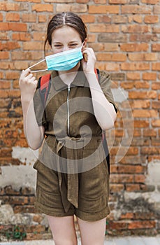 Schoolgirl poses against a brick wall in the backyard of the school, wearing a protective mask on her face from a coronavirus