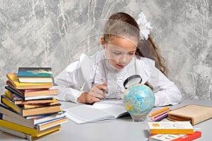 The schoolgirl in school uniform looking at a globe through a magnifying glass and writing in a notebook.