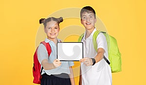 A schoolgirl and schoolboy with backpacks smilingly present a tablet with a white screen photo