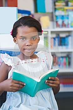 Schoolgirl sitting on chair and reading book in library