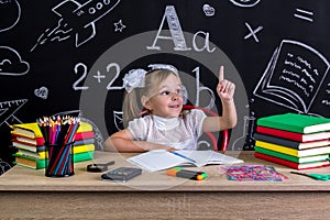 Schoolgirl sitting at the desk with books, school supplies, holding her pointing finger up. Coming up with the right