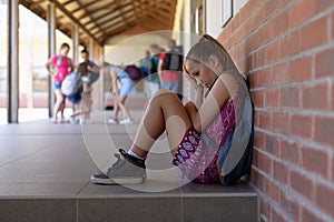 Schoolgirl sitting on the ground against a wall alone in the schoolyard at elementary school