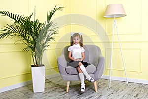 Schoolgirl sitting on the luxurious grey armchair in classic style in the room with the palm tree in the vase and a